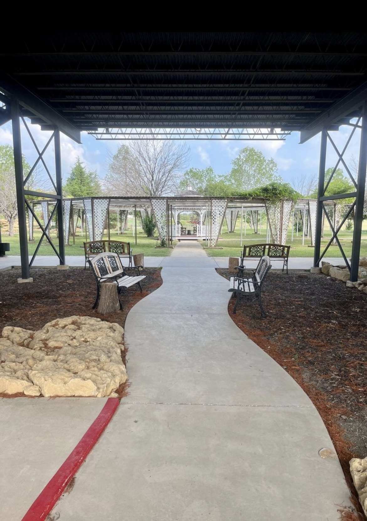 A curved concrete walk leads out from under the pavilion past benches and limestone rocks to white garden arbors.