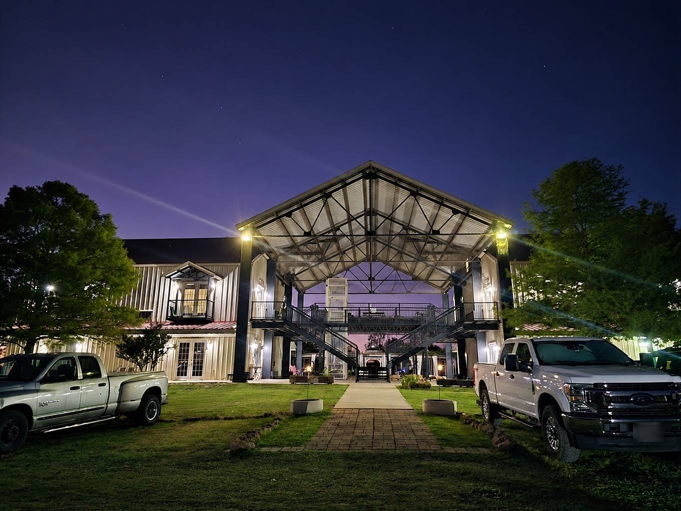 The two-story studio building at night, its big steel pavilion lit from inside, with a brick path across the lawn and two pickups parked out front.