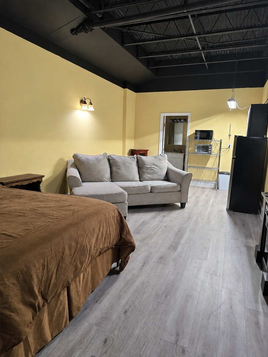 Studio 105: a bed with a brown spread, a grey sofa, wood-look floors and the kitchen shelving at the far end.