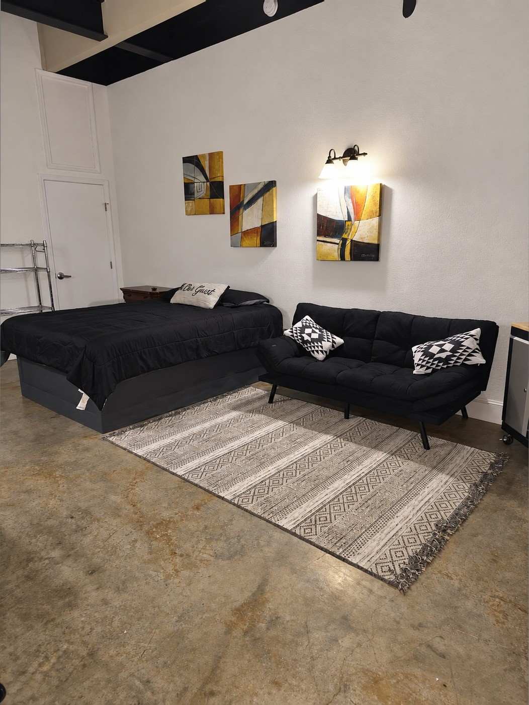 Studio 201: a bed with a black comforter, a black futon sofa on a striped rug, framed art and a wall lamp on white walls.