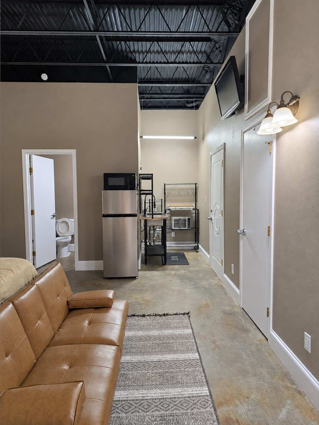 Studio 204 seen from the sofa: a tan leather futon, a runner rug, and a fridge, microwave and bathroom at the far end.
